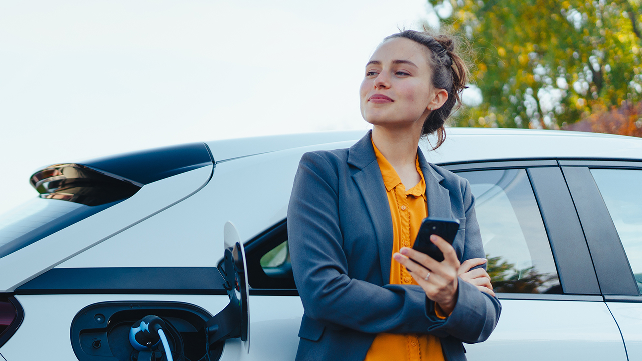 Woman standing next to an electric vehicle while holding a smartphone, with the car connected to a charging station.