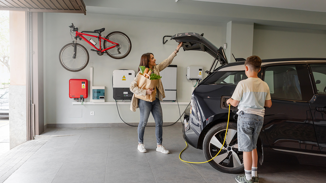 A woman unloading groceries from an electric car while a child connects the car to a home charging station.