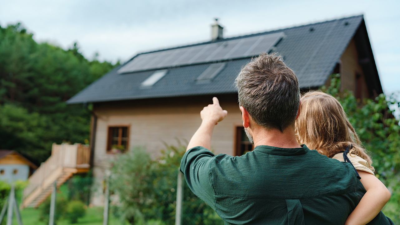 A father holding his child and pointing towards a house with solar panels on the roof.