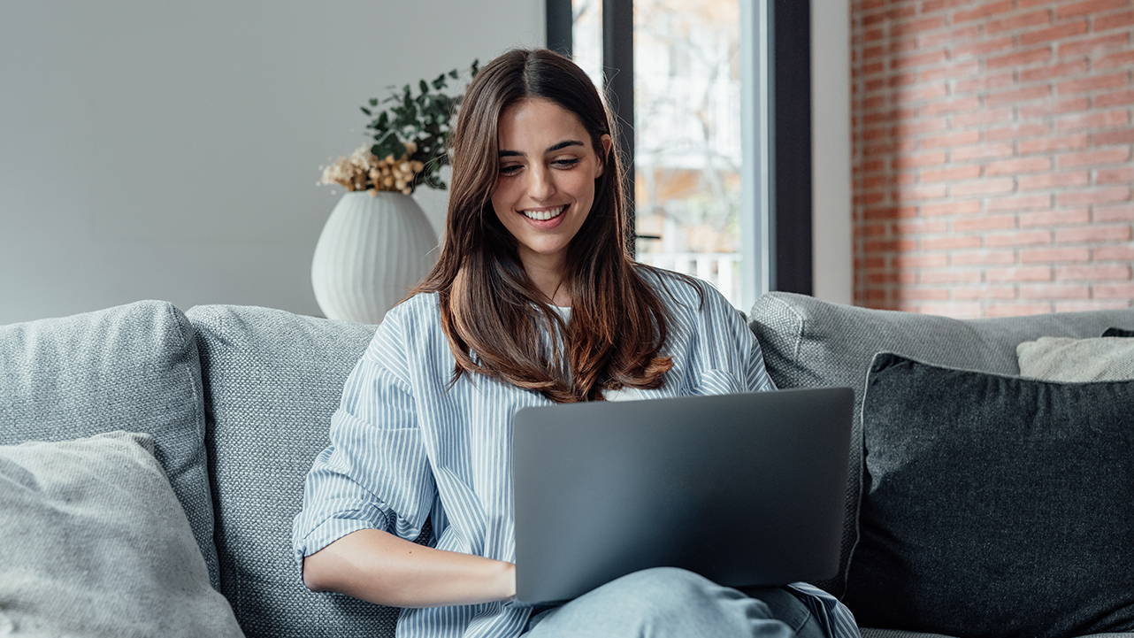 A smiling woman sitting on a sofa and working on her laptop in a bright, modern living room.