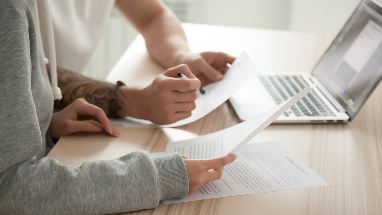 Two people reviewing documents at a desk with a laptop, comparing loans details.